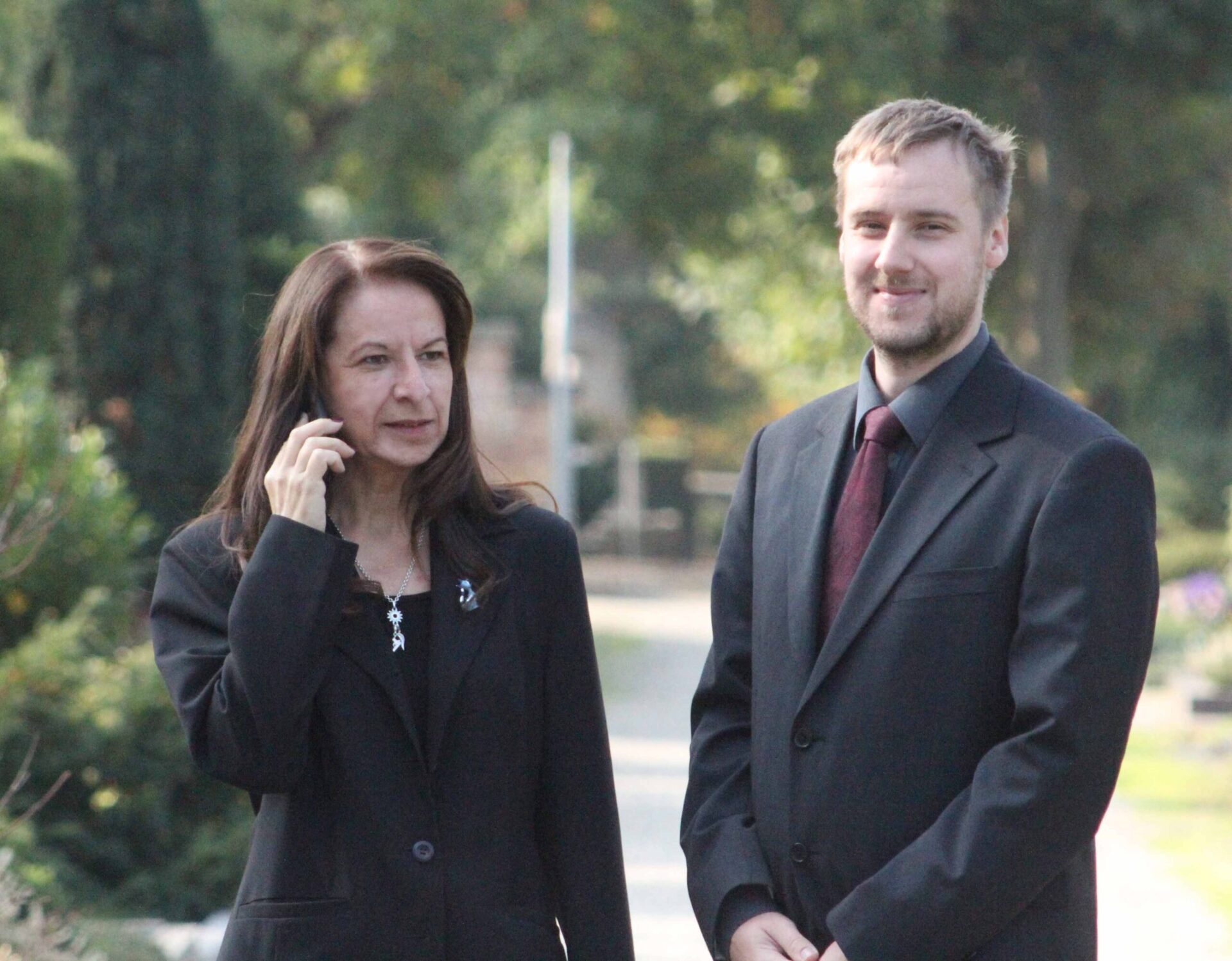 Zwei Personen in schwarzer Kleidung stehen nebeneinander im Freien, eine Frau telefoniert, ein Mann blickt lächelnd in die Kamera.